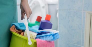 Mulher segurando um balde com vários produtos de limpeza. Dentre eles estão os tipos de detergente.
