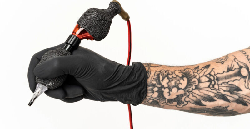 Braço tatuado com luva preta segurando máquina representando limpeza em estúdio de tatuagem.