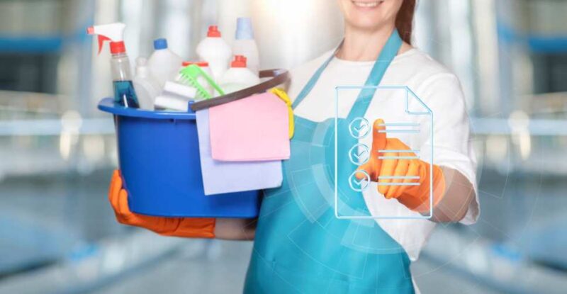 Pessoa com luvas e avental segura balde azul cheio de produtos, representando checklist de limpeza.