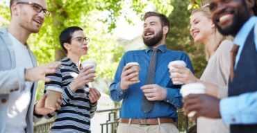 Saiba como organizar um coffee break ao ar livre Grupo de colegas sorri e conversa em um coffee break ao ar livre com copos de café.